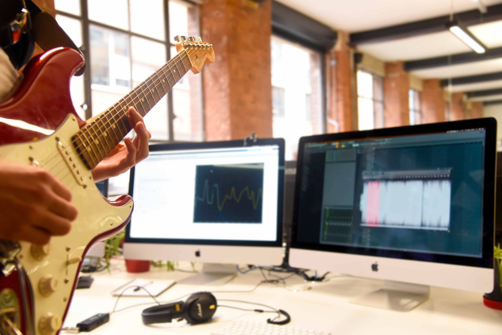 Playing an electric guitar at a desk beside two monitors showing audio software